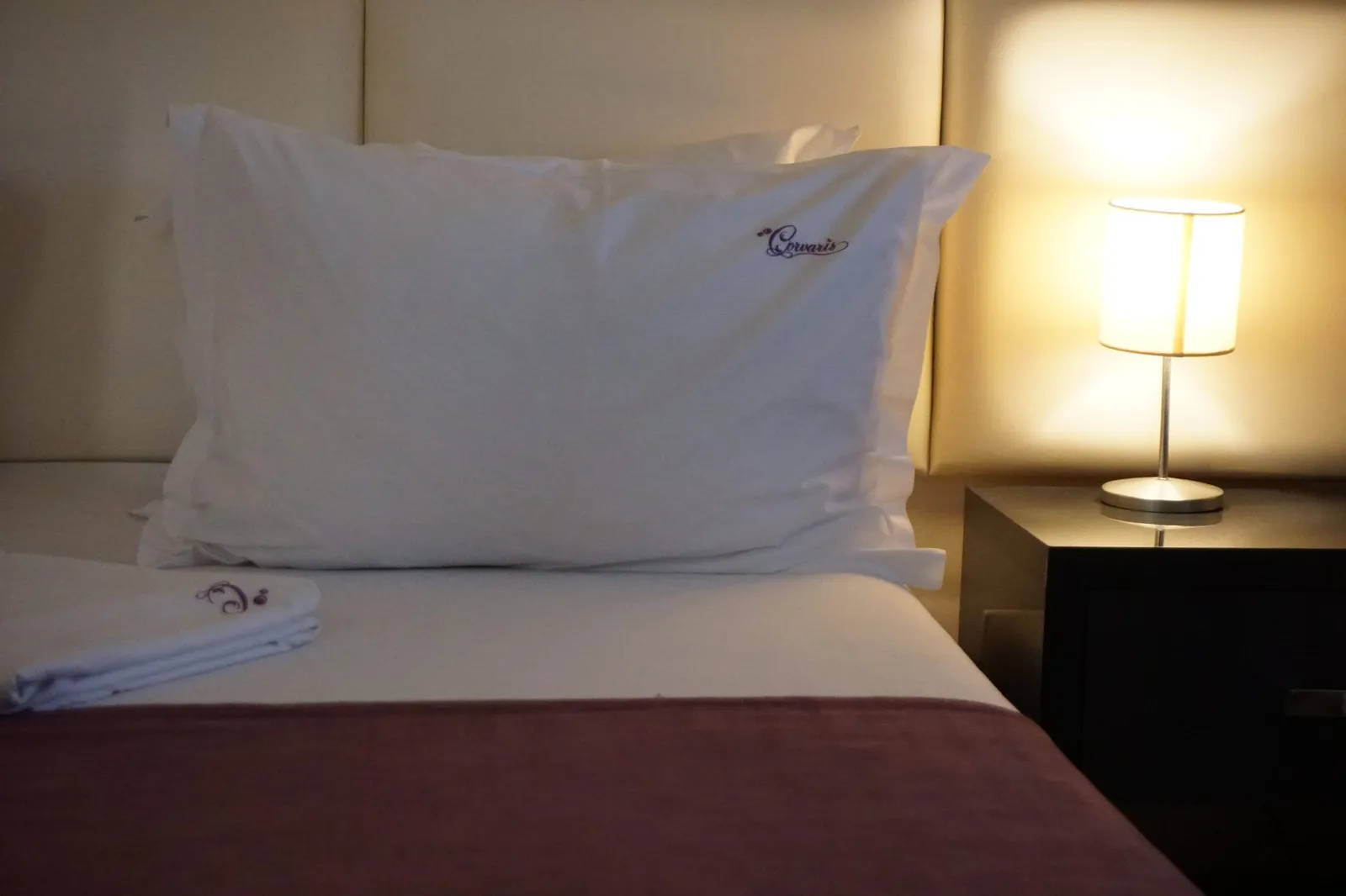 A pillow embroidered with the Corvaris logo, next to the lit bedside lamp