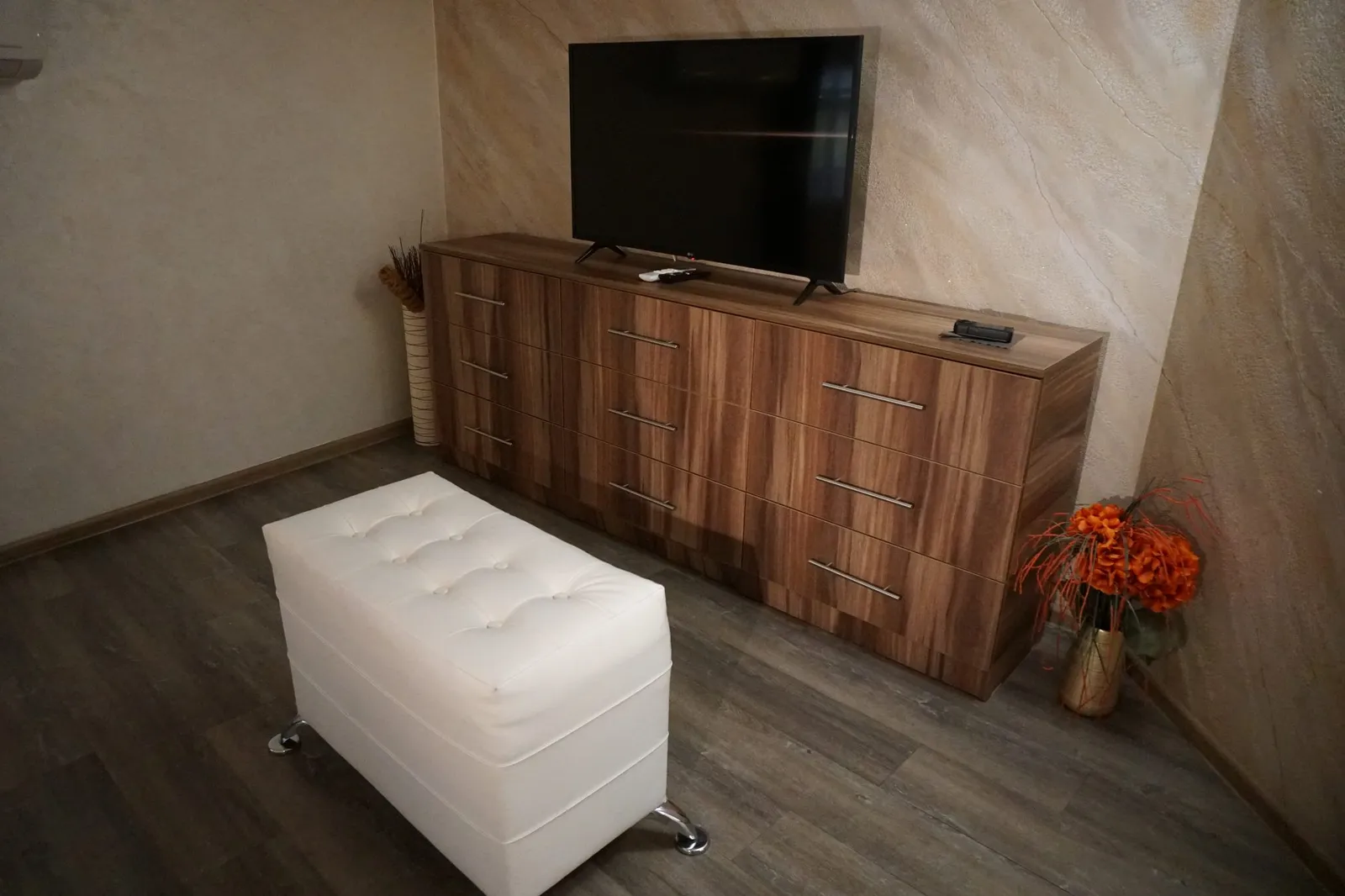The chest of drawers with a TV and the tufted ottoman in the bedroom